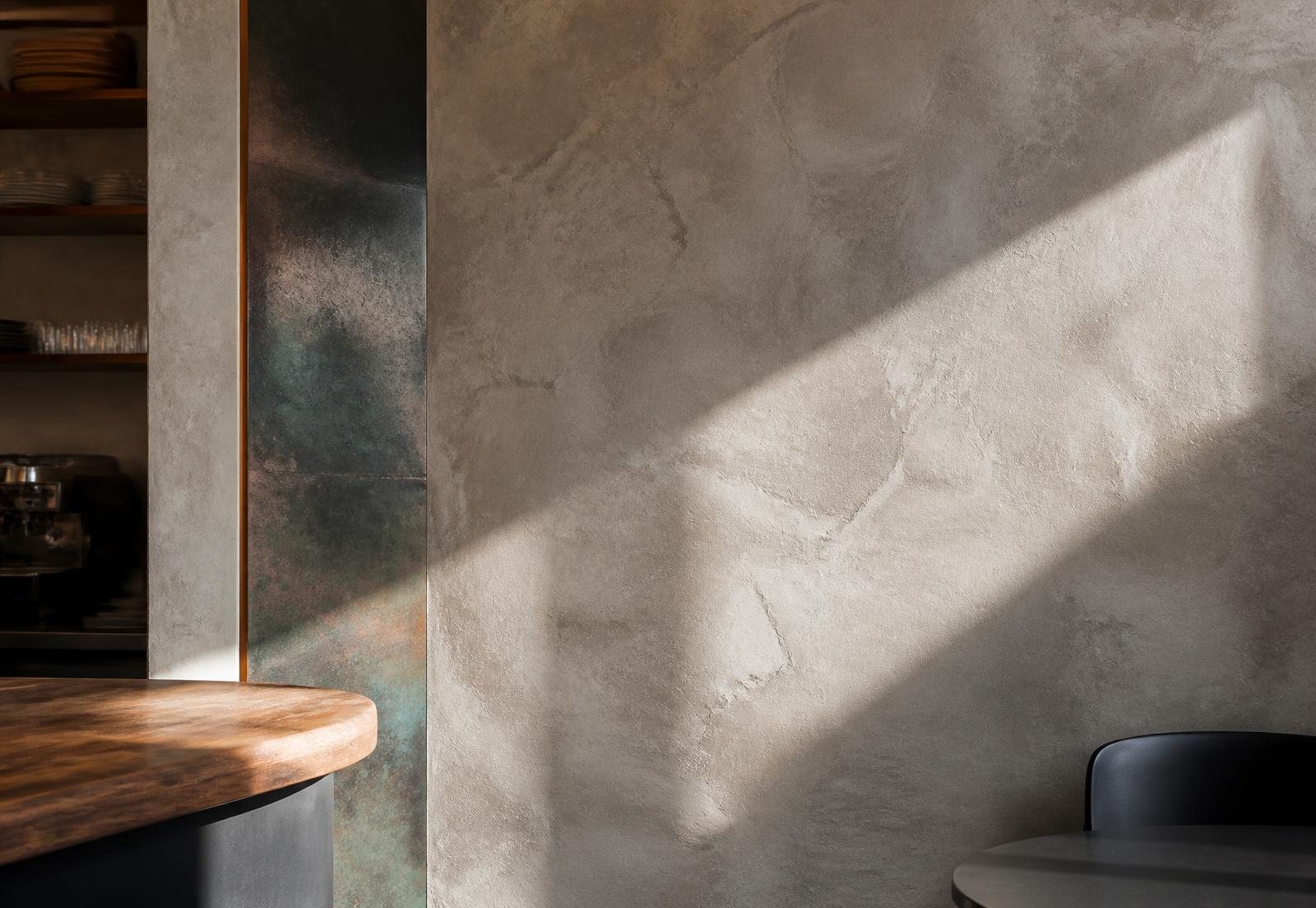 Interior detail of a café: plaster wall, oxidised metal panel and a timber counter edge in raking daylight.